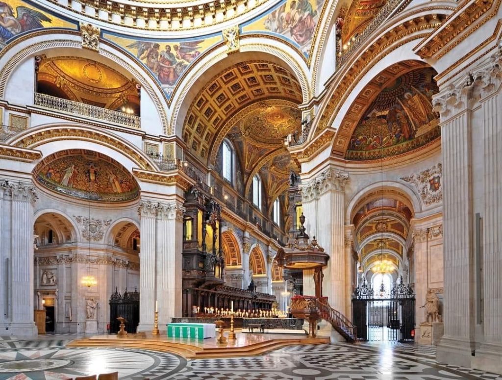 Inside of St. Paul's Cathedral. Soaring ceilings with elaborate paintings and gold gilding. The great hall is surrounded by high stone arches with carved columns leading to openings. The floor is a gleaming black and white checked tile/marble design. A raised wooden dais is in the middle of the room with large candlesticks and an altar.