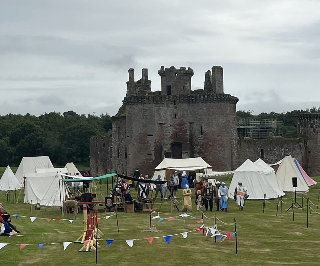Castle in the background with one large round turret and four chimneys on either side. In the foreground are tents - carnival like - with people in period costumes walking around the green grassy lawn.