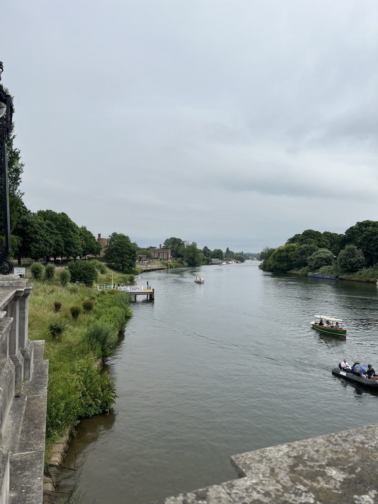View from the bridge walking into Hampton Court of the River Thames. There are people swimming and boating on the river. River banks are filled with trees and a glimpse of the palace.