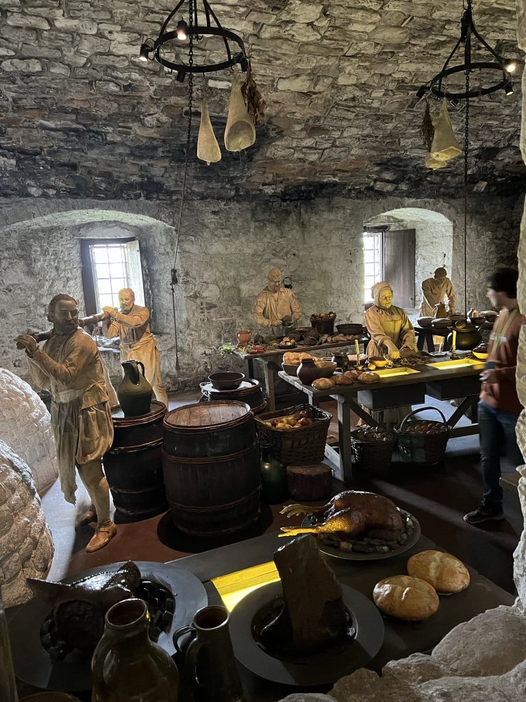 The kitchen area at Stirling Castle. There are life-sized figures to represent the busy kitchen area. Two figures of men are carrying a "deer". There are barrels of wind and spices next to a table filled with bread and figures of ladies behind the table cooking. Suspended form the rock ceiling are bags of spices. Two windows provide light for the area.