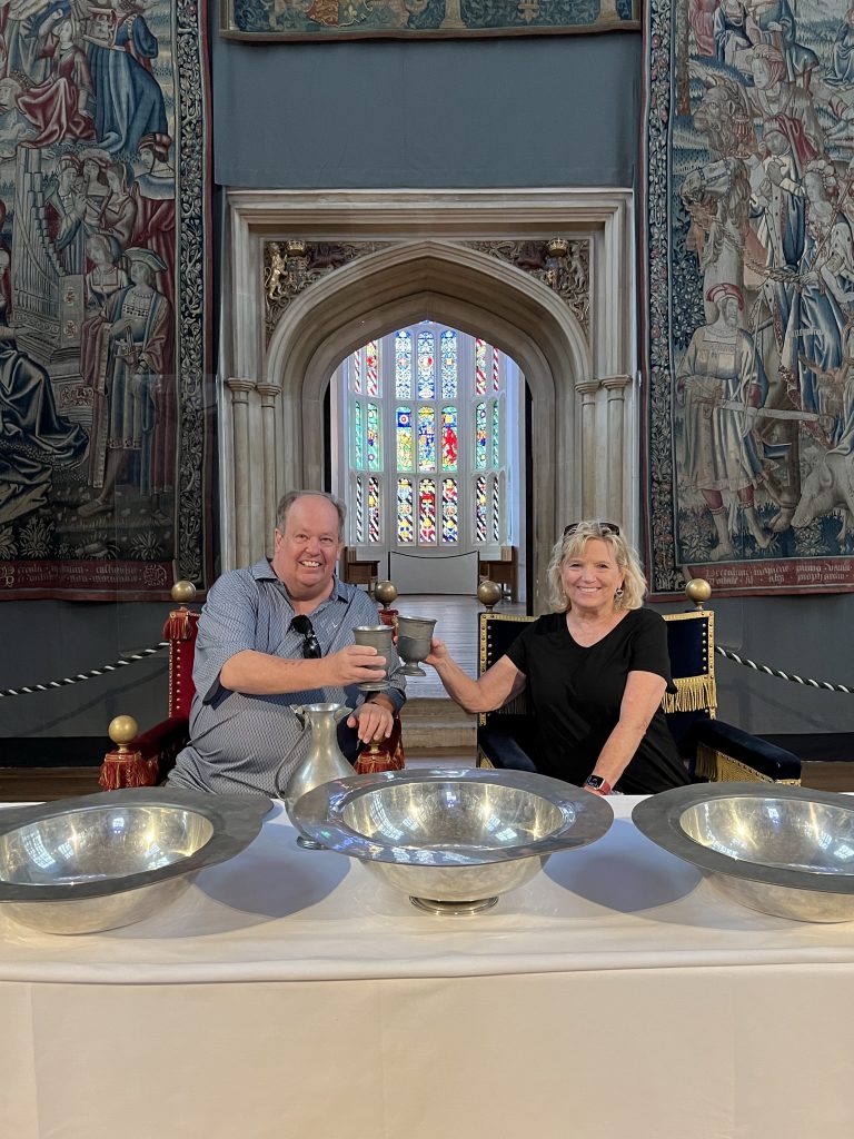 Kenny and Kay sitting at the King's table in the great hall at Hampton Court. The table is set with large silver platters and bowls. Heavy tapestries surround the walls. Kenny and Kay are toasting each other with silver chalices