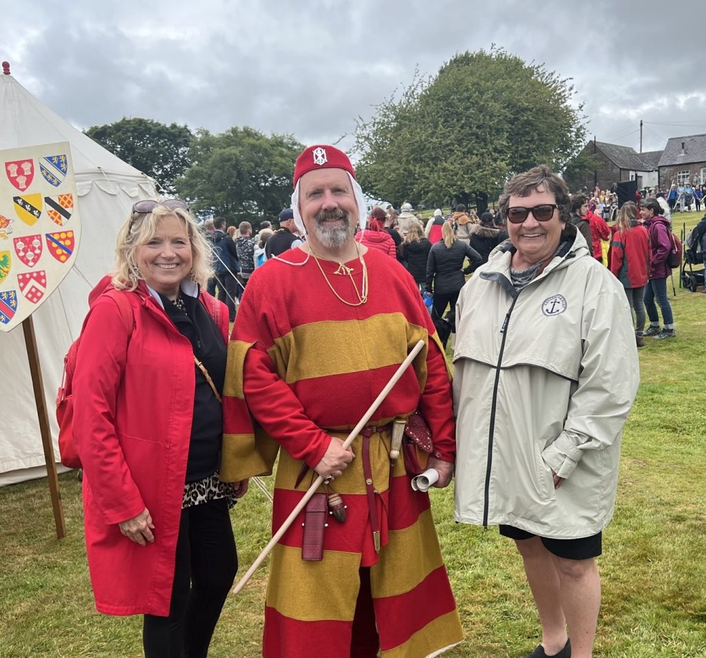 The kings herald dressed in a yellow and red tunic with a red hat standing between Kay and Kathy. The herald is holding a rolled scroll.