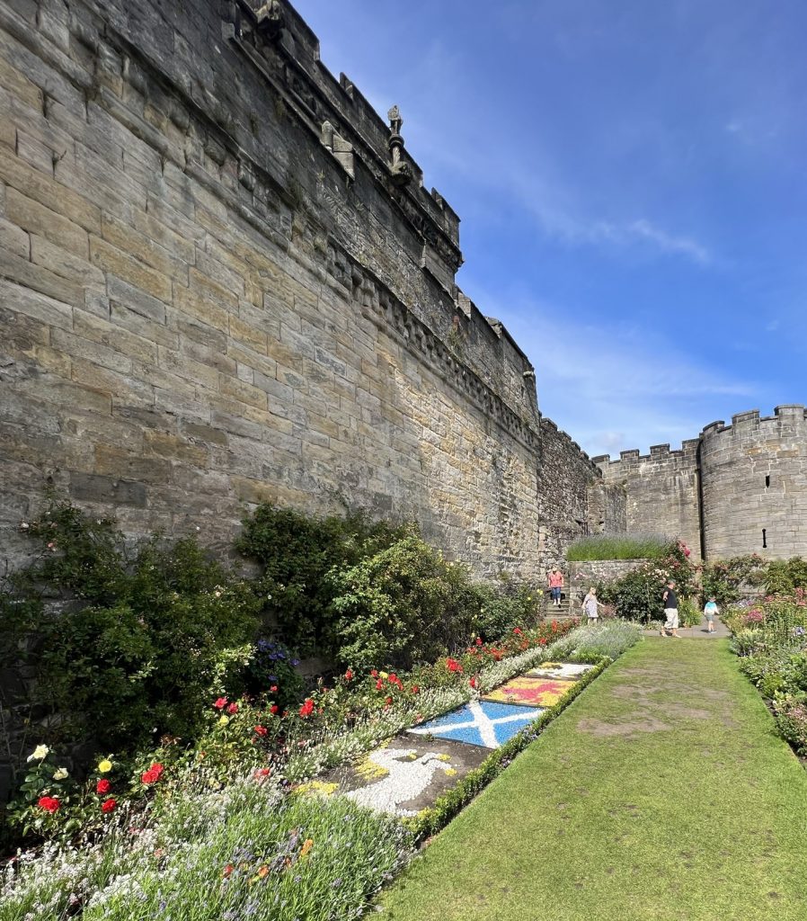 Near the castle wall there is a terrace area of the garden. This terrace has a flower bed that has three rock garden areas. One rock garden is the blue and white Scottish flag, one is the yellow and red Scottish Lion emblem and one is a unicorn, the emblem of Scotland. These rock garden areas are bordered by flowers and greenery next to the castle walls.