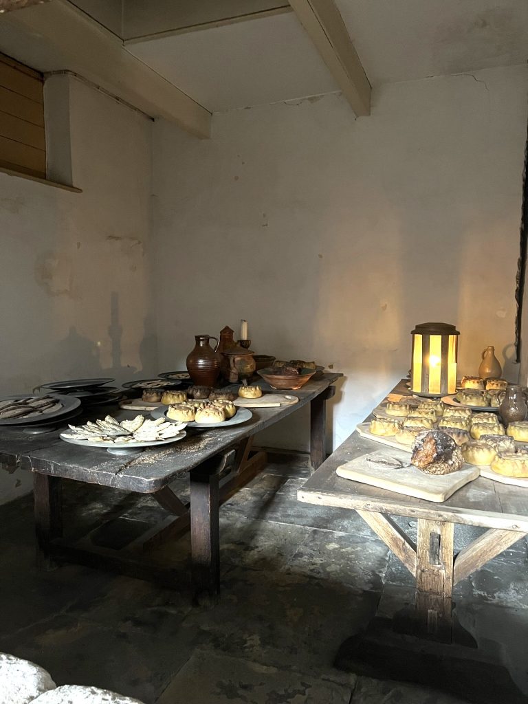 Two tables with food for the banquet. both tables are filled with breads, scones, rolls, baked meat pies, candles, jugs and platters.