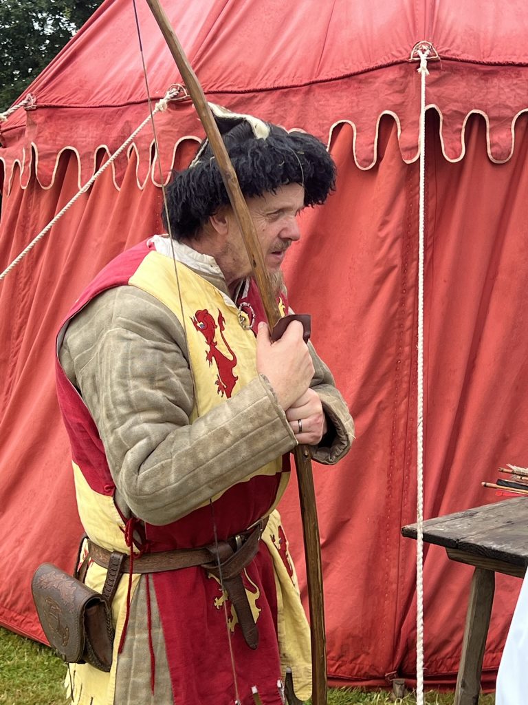 A bowman holding his large bow. He is dressed in a yellow and red tunic with the Scottish lion and unicorn crests.