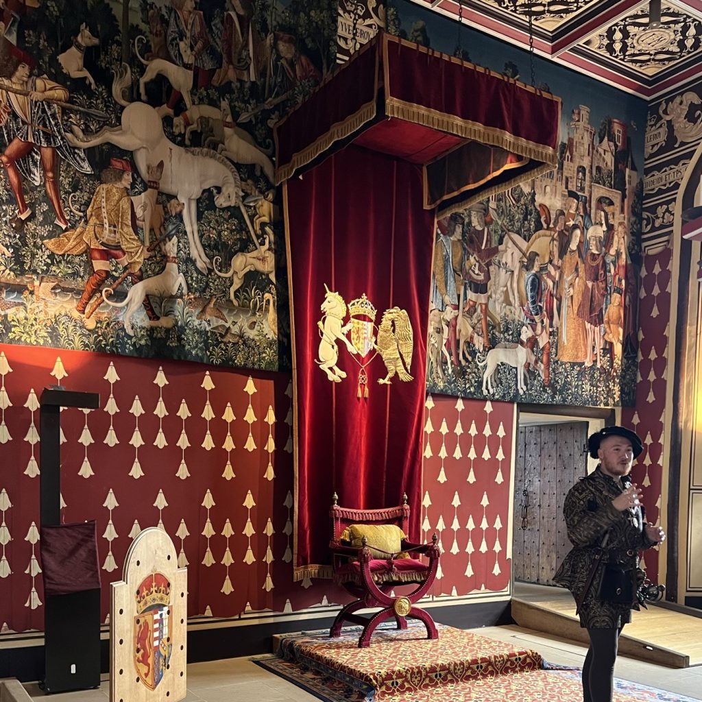 The throne chair sits on a small raised platform. The chair has deep red cushions and a yellow pillow. Above the throne is a velvet hanging with the unicorn, the crown, and the eagle representing Scotland. The bottom half of the wall has dark red wallpaper with gold decorations. The top half of the wall is covered with several panels of the tapestries.
