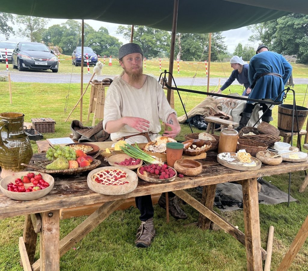 A kitchen worker sitting at a table filled with medieval type food. There are fruits and vegetables, meat pies, rasberries, green onions, and cheese.