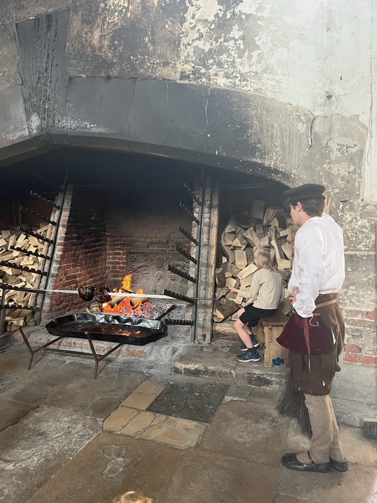 The grand fireplace with a roasting pan to catch the drippings from the meats being cooked. Above the fire, there are 8 rotisserie arms with slits for 8 rotating spits for a grand total of 64 types of meat to be cooked at the same time. Today has one chicken that is being roasted by a young boy (probably a tourist) and watched by a young man in medieval dress. There are two 5 foot stacks of firewood on either side of the fireplace.
