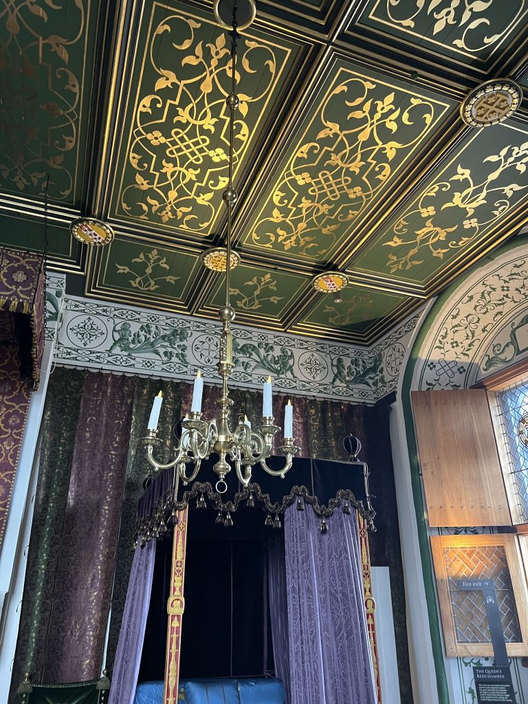 The queens ceremonial bedchamber with an elaborate bed with purple drapes and a velvet dark purple canopy. The canopy has silver details and scalloped edges. Behind the bed ore more purple drapes. The ceiling is elaborately painted in greens and whites.