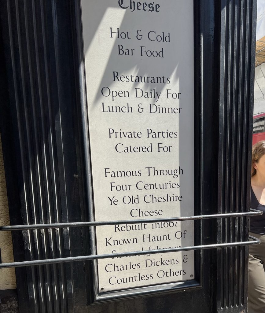 Image of the sign in front of the Ye Old Cheshire Cheese Pub. Sign reads: Hot & Cold Bar Food, Restaurants Open Daily For Lunch & Dinner. Private Parties Catered For Famous Through Four Centuries. Ye Old Chesire Cheese Rebuilt in 1667. Known Haunt of Samuel Johnson, Charles Dickens & Countless Others