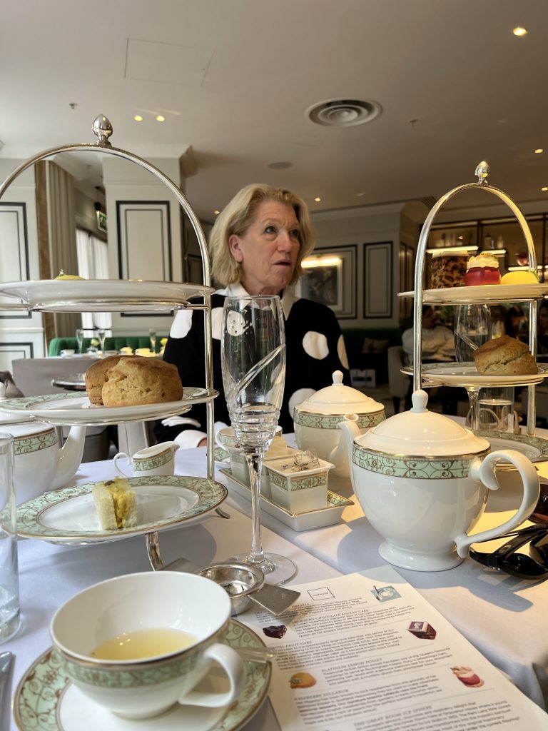 High Tea celebration with champagne glasses (empty), tea pots, tea cups, and two three tiered sandwich and pastry trays. The first tier was assorted finger sandwiches, the second tier was scones, and the third was pastries and chocolates. Jodi is sitting across the table.