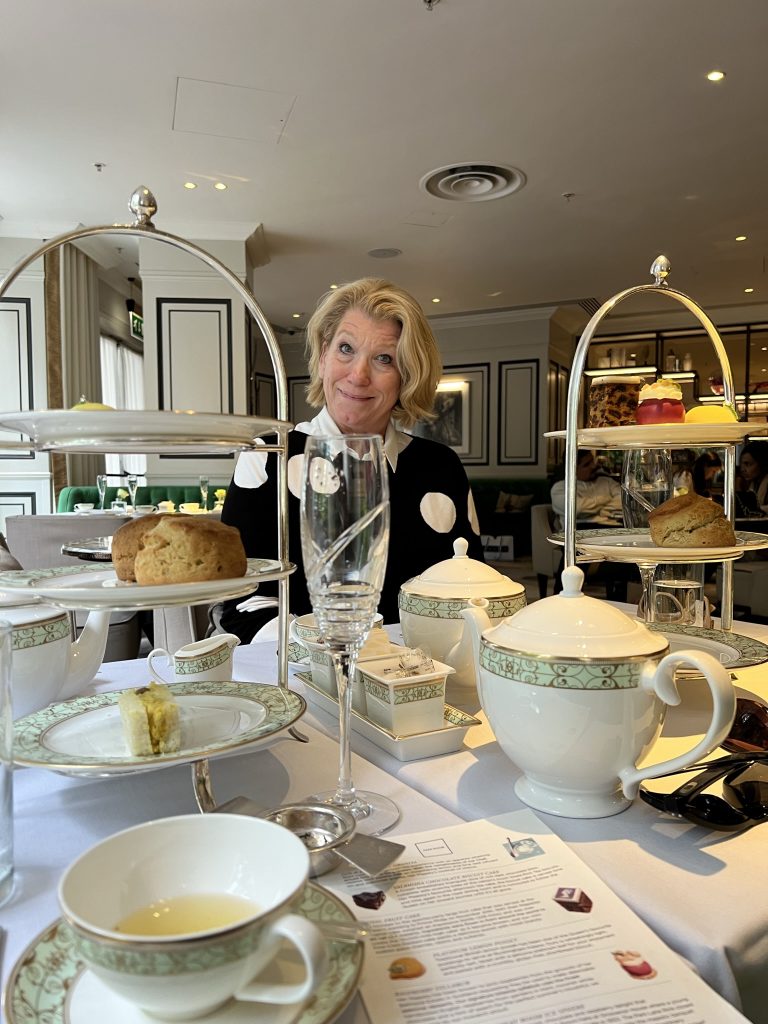 High Tea celebration with champagne glasses (empty), tea pots, tea cups, and two three tiered sandwich and pastry trays. The first tier was assorted finger sandwiches, the second tier was scones, and the third was pastries and chocolates. Jodi is sitting across the table.