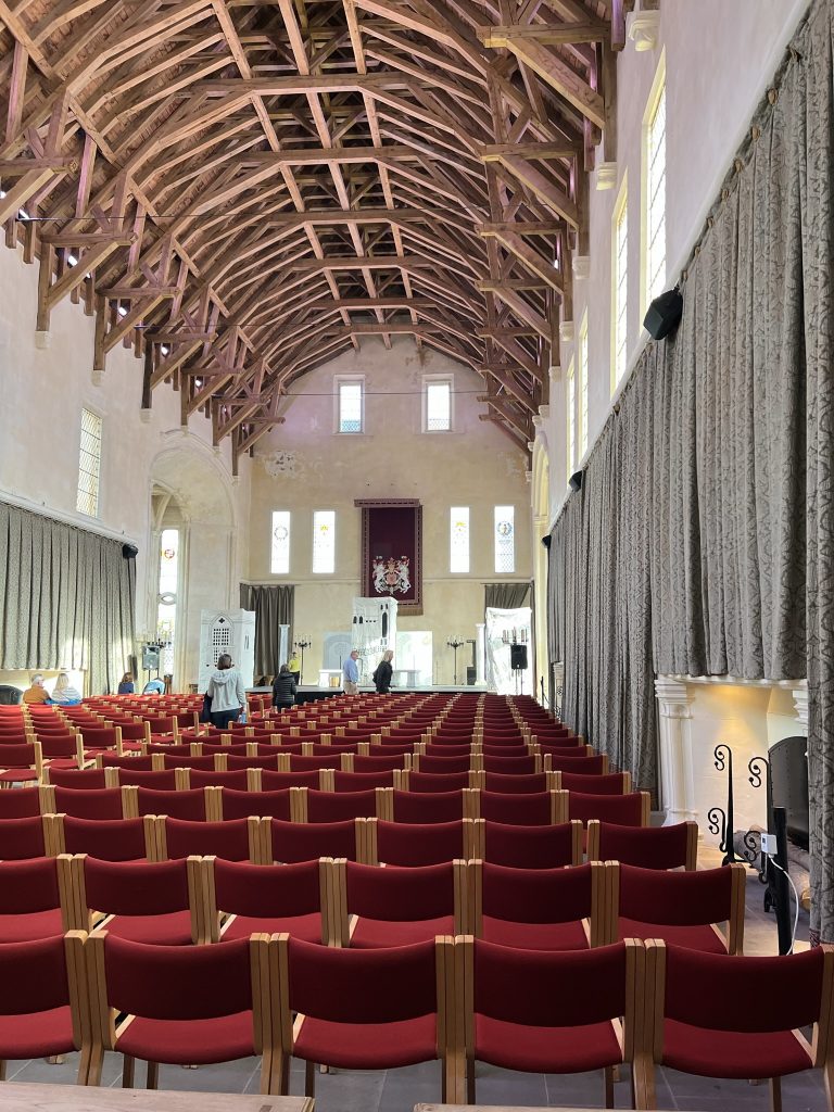 The Great Hall at Stirling Castle.  This is the largest great hall in Scotland.  The room is long and narrow with walls painted a light white.  The ceiling is magnificent with wooden beams that appear to be like a ship's hull.  Down from the ceiling is a row of windows then dark green curtains line the walls.  This room was set for a play so it is filled with red chairs.  