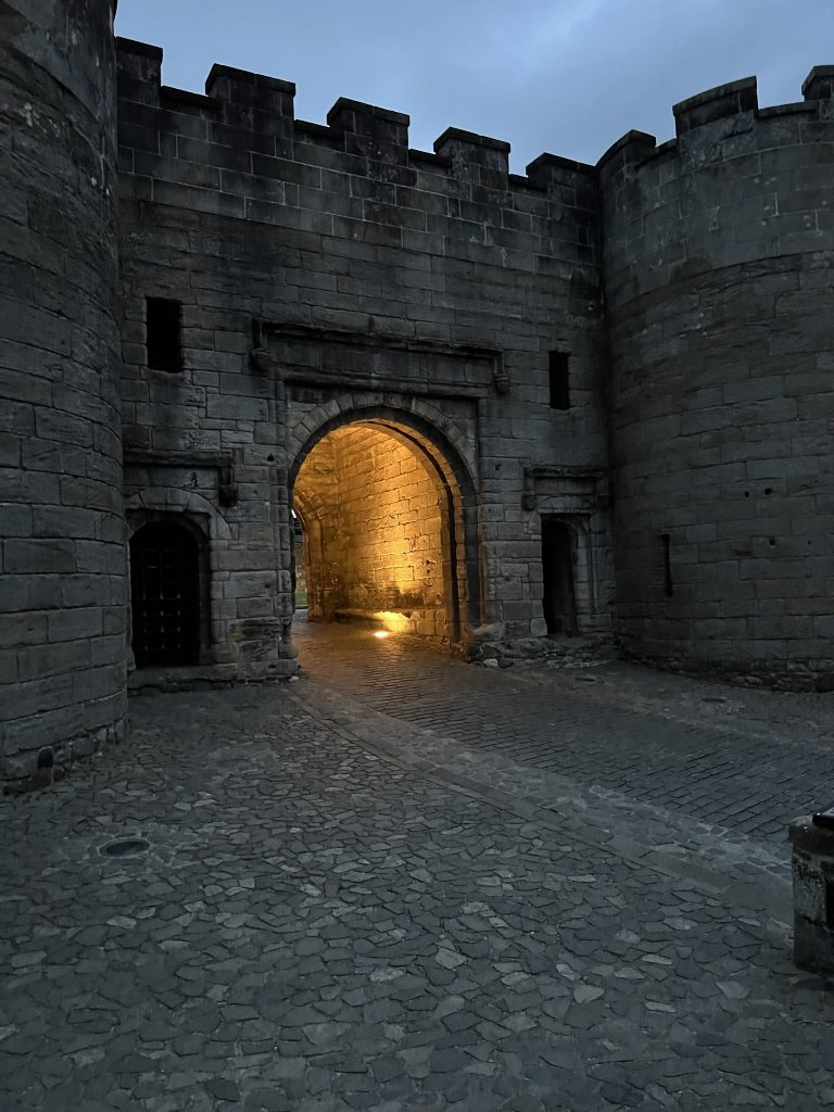 The castle entrance at night. The archway is lit up from within the castle and the two round towers on each side are visible.