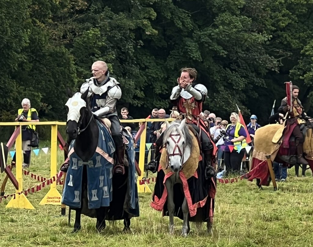 Two knights riding their horses at the end of the joust. One knight is clapping towards the crowd.
