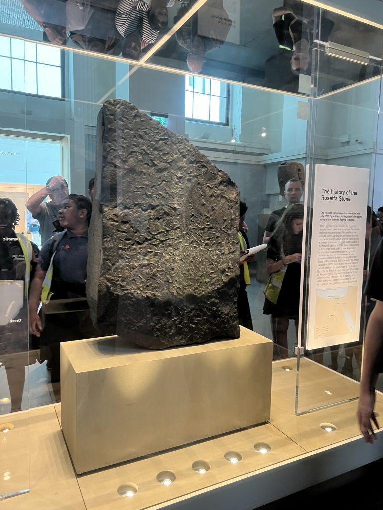 Image of a large slab of stone enclosed in a glass protective case. On one side the stone has inscriptions in hieroglyphics, Demoitic scripts, and Ancient Greek.