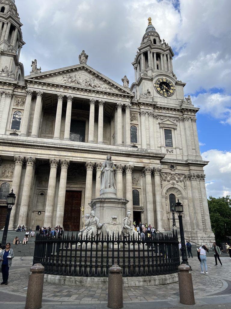 Front of St. Paul's Cathedral. Two towering columns - one with a large clock face - flank the center of the building. Two tiers with three sets of two columns on the top level and five sets of two columns on the bottom level. In the courtyard is a large statute of Queen Victoria with a black metal fence surrounding it. Tourist are milling around the front of the cathedral and sitting on the stairs leading up to the massive doors.