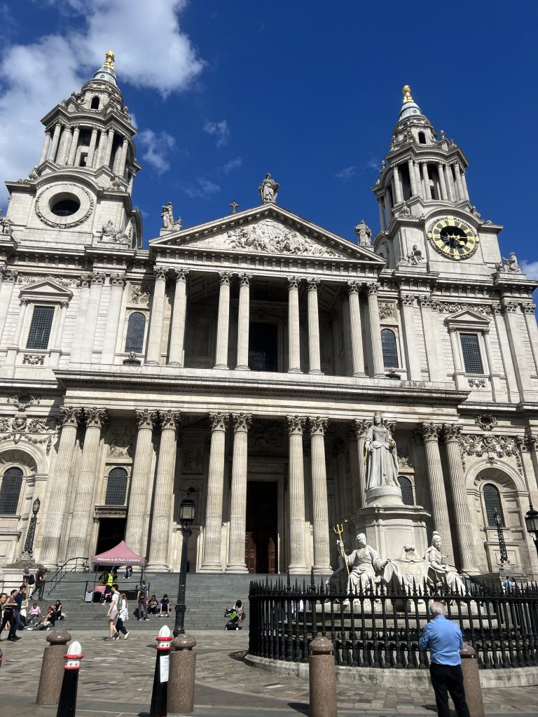 Front of St. Paul's Cathedral. Two towering columns - one with a large clock face - flank the center of the building. Two tiers with three sets of two columns on the top level and five sets of two columns on the bottom level. In the courtyard is a large statute of Queen Victoria with a black metal fence surrounding it. Tourist are milling around the front of the cathedral and sitting on the stairs leading up to the massive doors.