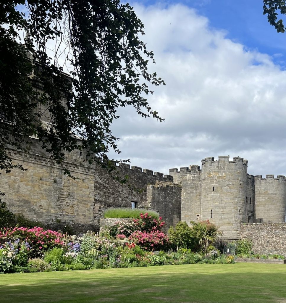 Queen Anne Garden inside the walls of Stirling Castle. A green grass lawn area leads up to a wide flower bed filled with flowers and greenery of white, pink, bule, purple, yellow and red flowers.  Behind the flowers are the castle walls of gray and yellowish stones with the iconic round tower. 