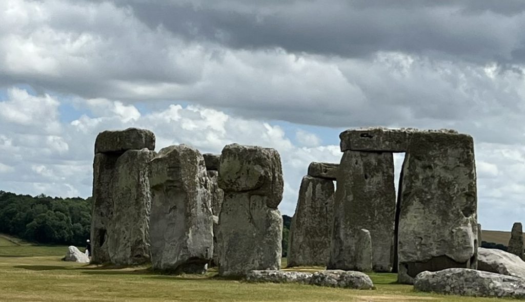 Image of the edge of the Stonehenge stone circle. Three upright stones without a lentil or horizontal stone on top, then two upright stones with a lentil. Stones are weathered and gray.