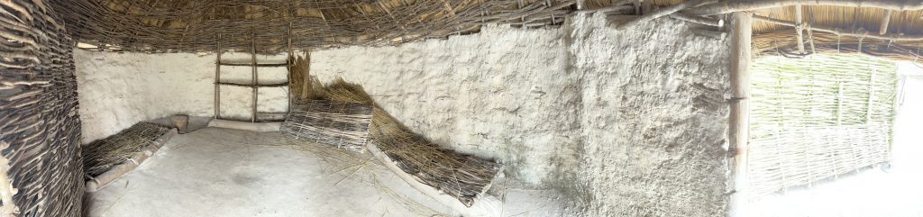 This panoramic view shows the sleeping space next to the cooking space with another sleeping space with extra woven matts. The circular hut has a woven straw door like opening.