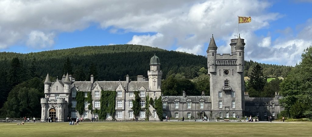 Balmoral Castle from the front.  Balmoral is a gray stoned castle with four distinct building areas.  The far end has a wooden door leading to a small enclosed garage like area.  This has two small round towers on either side.  The building continues with seven sets of windows, each set three high.  There two small turrets on one end and a larger tower with a dome on the other end of this second section.  Connecting this building to a larger tower is a smaller area with four sets of two window.  Third, there  is a large tower with four turrets and a Scottish flag flying from one of the turrets.   Adjoining this tower is another section that is lower.  This area is an enclosed courtyard area where the cars can pull up to load and unload people without being seen.  The castle sits in front of a large Highland mountain filled with trees.  in Front of the castle is a large green area bordered with trees and gardens.  
