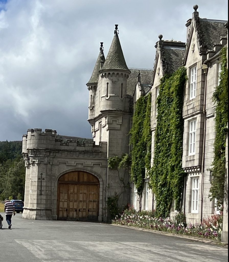 The front garage area from the side showing an elaborate wooden door that opens to an enclosed garage-like area. There are flowers beds in the front of the castle and ivy growing up the front two sections that do not have windows.  Tow small turrets of the garage and two large turrets of the house can be seen. 