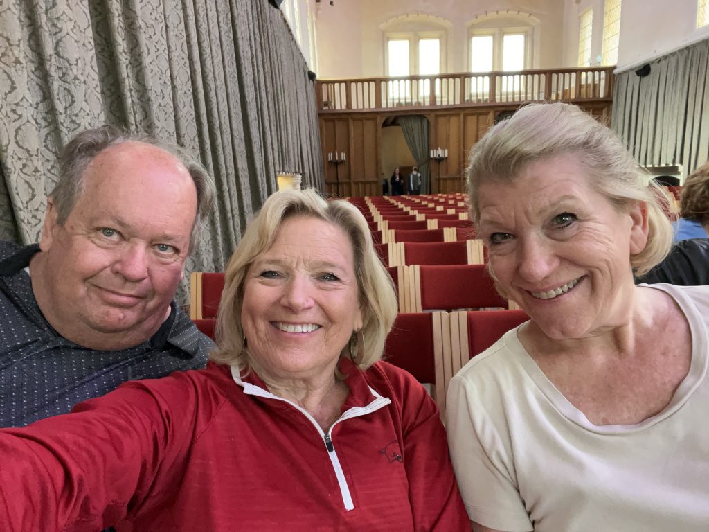 Kenny, Kay and Jodi sitting in the great hall at Stirling Castle.  There are rows red chairs lining the hall.  In the background is the wood decorative area that separated the hall from the entry.  The green curtain-like wall hangings line the walls. 