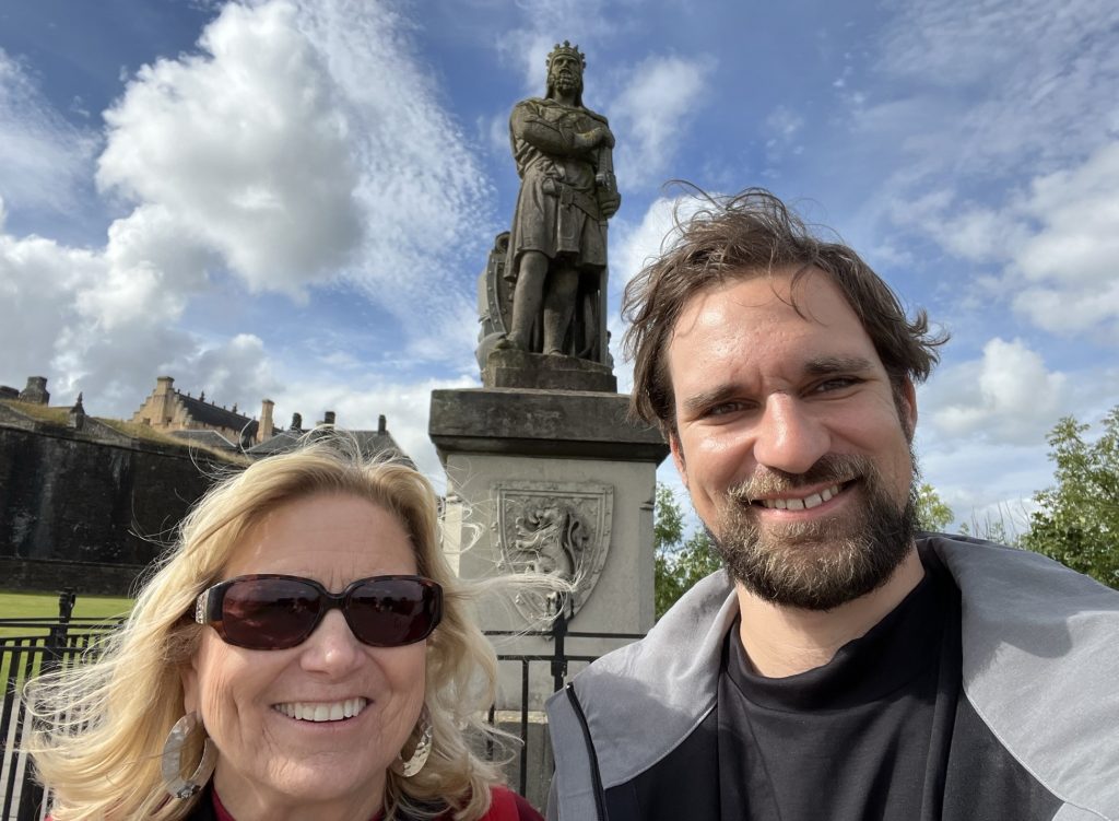 Kay and Will smiling while standing in front of the King Robert the Bruce statue. In the background is the Great Hall.  There are blue skies with white wispy clouds and lots of sunshine. 