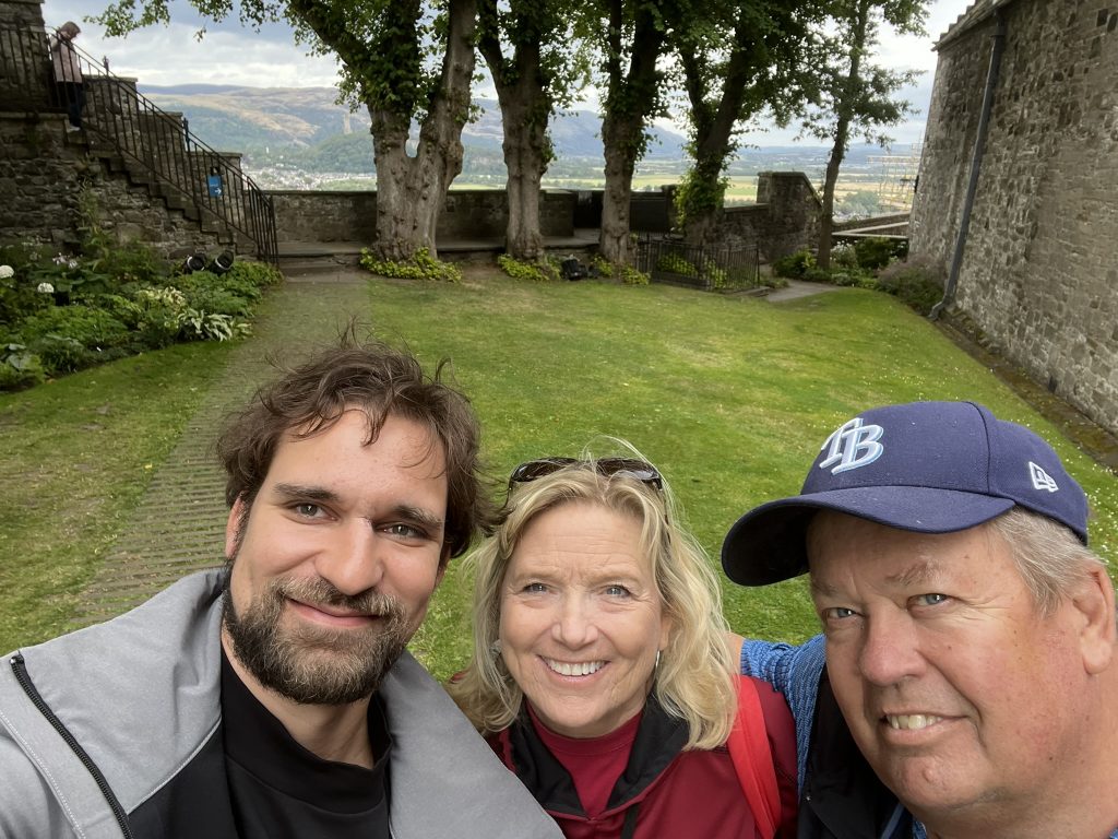 Will, Kay and Kenny in the Douglas Garden.  There is a tall rock wall on one side with a lower wall and free trees in the background.  Beyond the trees are the hills of the Highlands. 