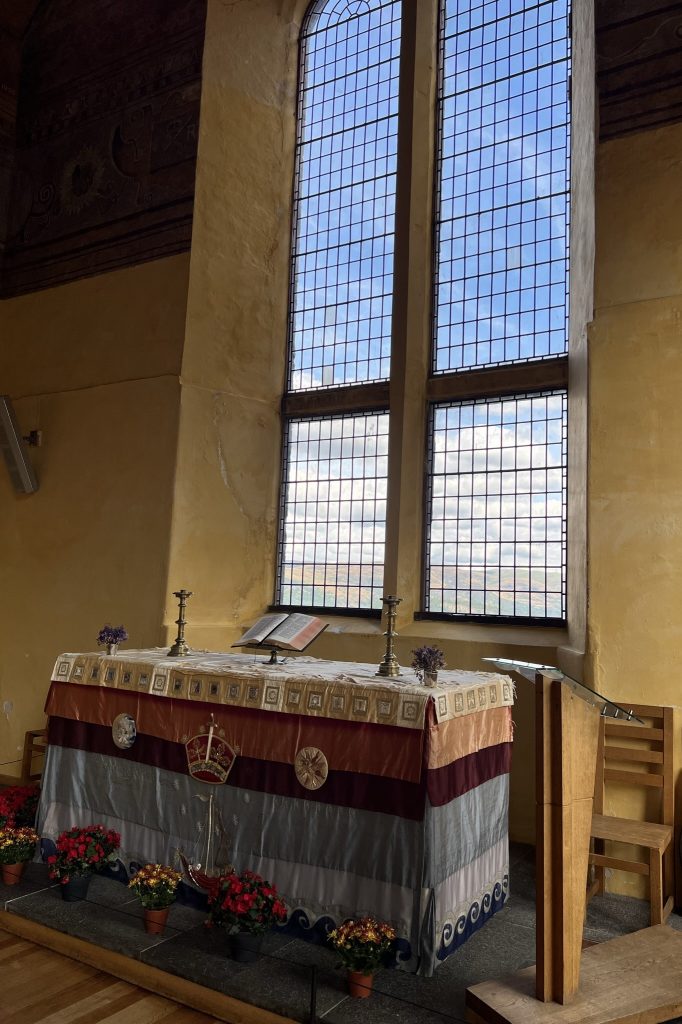Altar in the Chapel Royale.  The altar is draped with a multi-colored fabric. The bottom is a silver scalloped edge, then a light silvery blue stripe, a dark maroon stripe, and a red satin type fabric.  The top is a gold and white patterned overlay.  On the altar are two candle sticks and a bible on a stand.  Behind the altar is a large two-story high arched window. The walls are painted a dark "kings gold" color. 